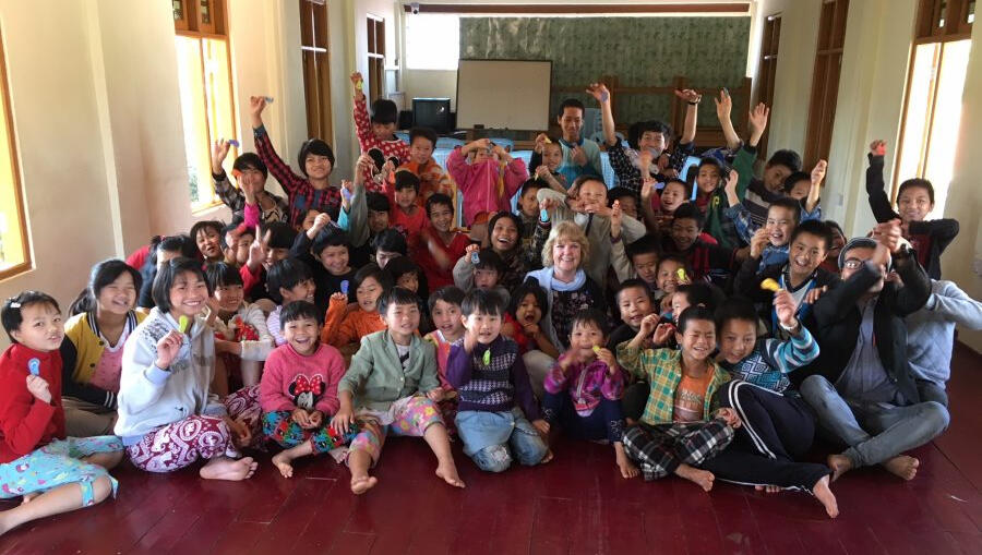 Lyn sits on the floor in the middle of a group of children everyone is smiling; some are waving their arms.